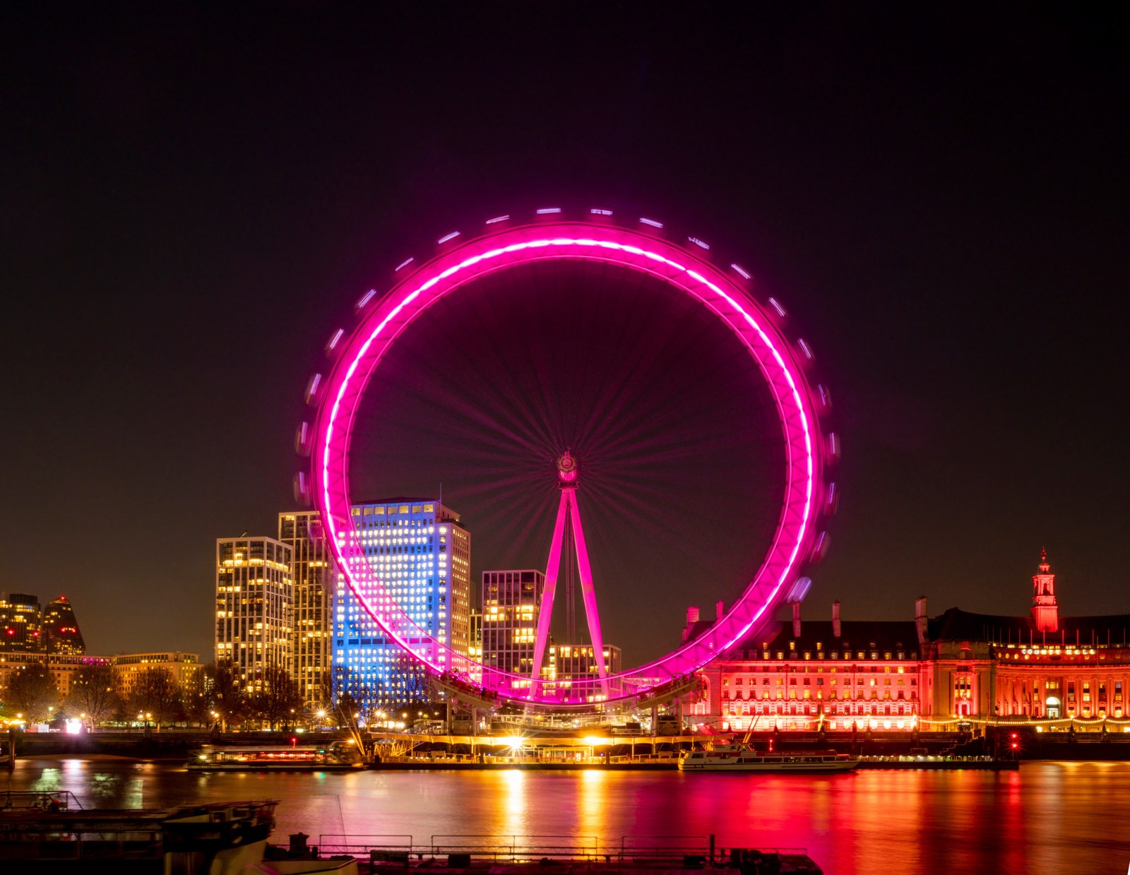 The London Eye glowing pink at night above colourful reflections on the River Thames.